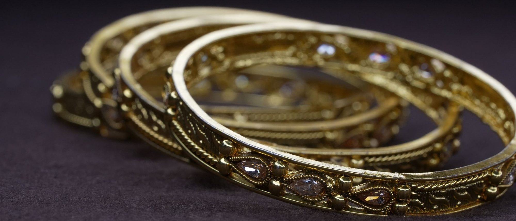 Three gold bangles with fine filigree and small stones resting on a dark cloth