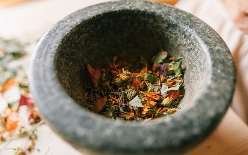 A granite mortar filled with dried herbs and petals on a pale wooden table