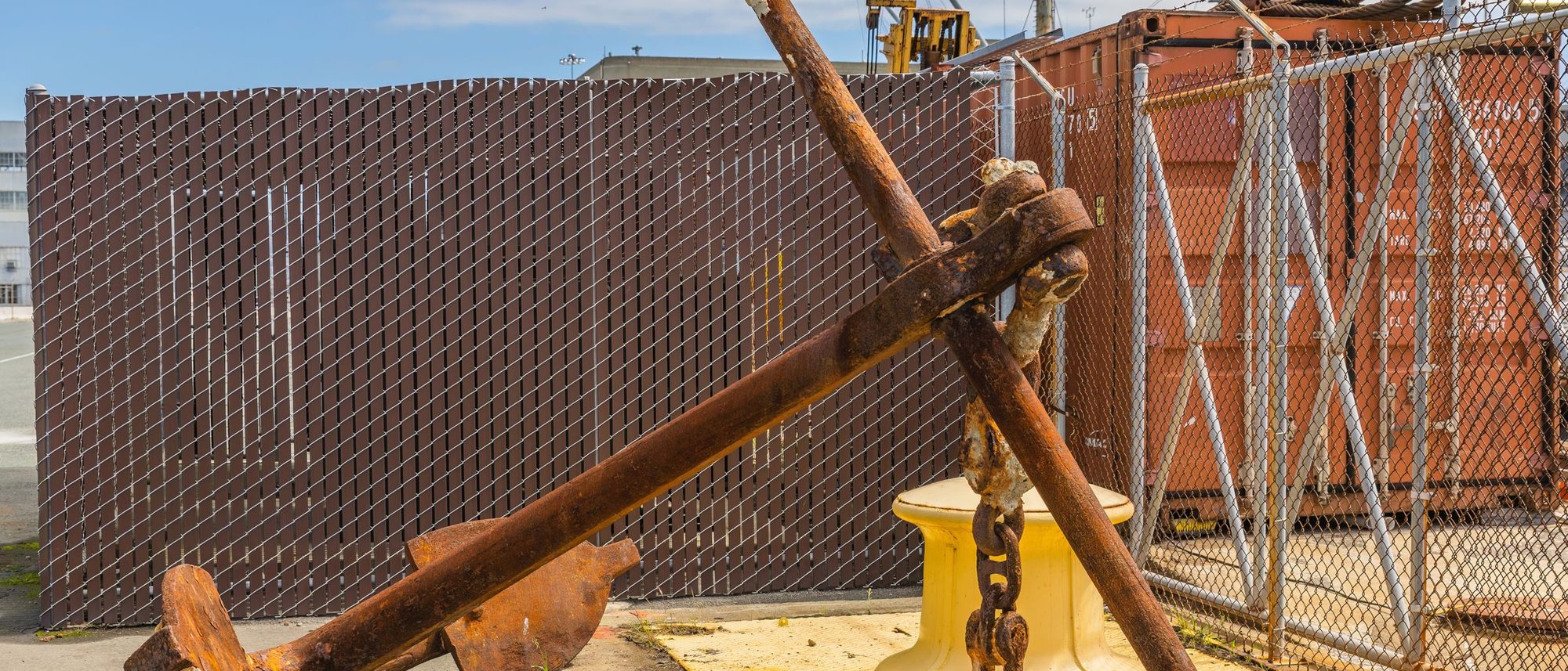 A large rusty ship’s anchor rests on a quay beside stacked containers.
