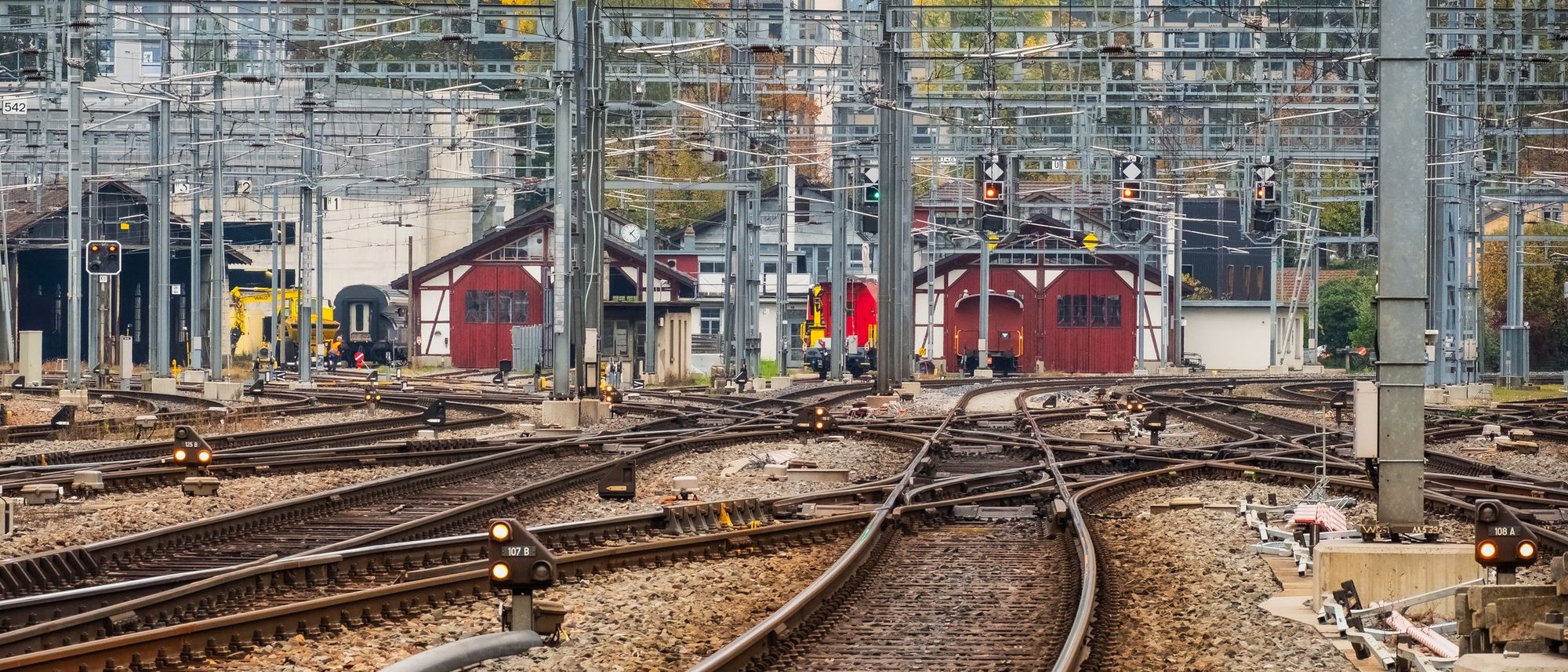 Several railway tracks running side by side past signals and buildings in daylight.