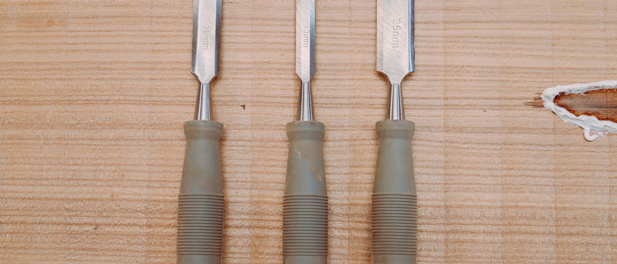 Three metal chisels laid side by side on a wooden surface, seen from above.
