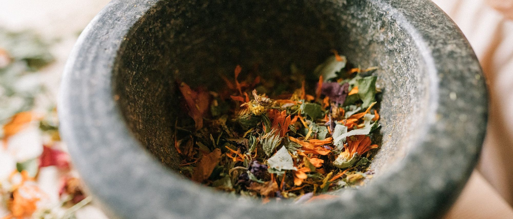 A granite mortar filled with dried herbs and petals on a pale wooden table
