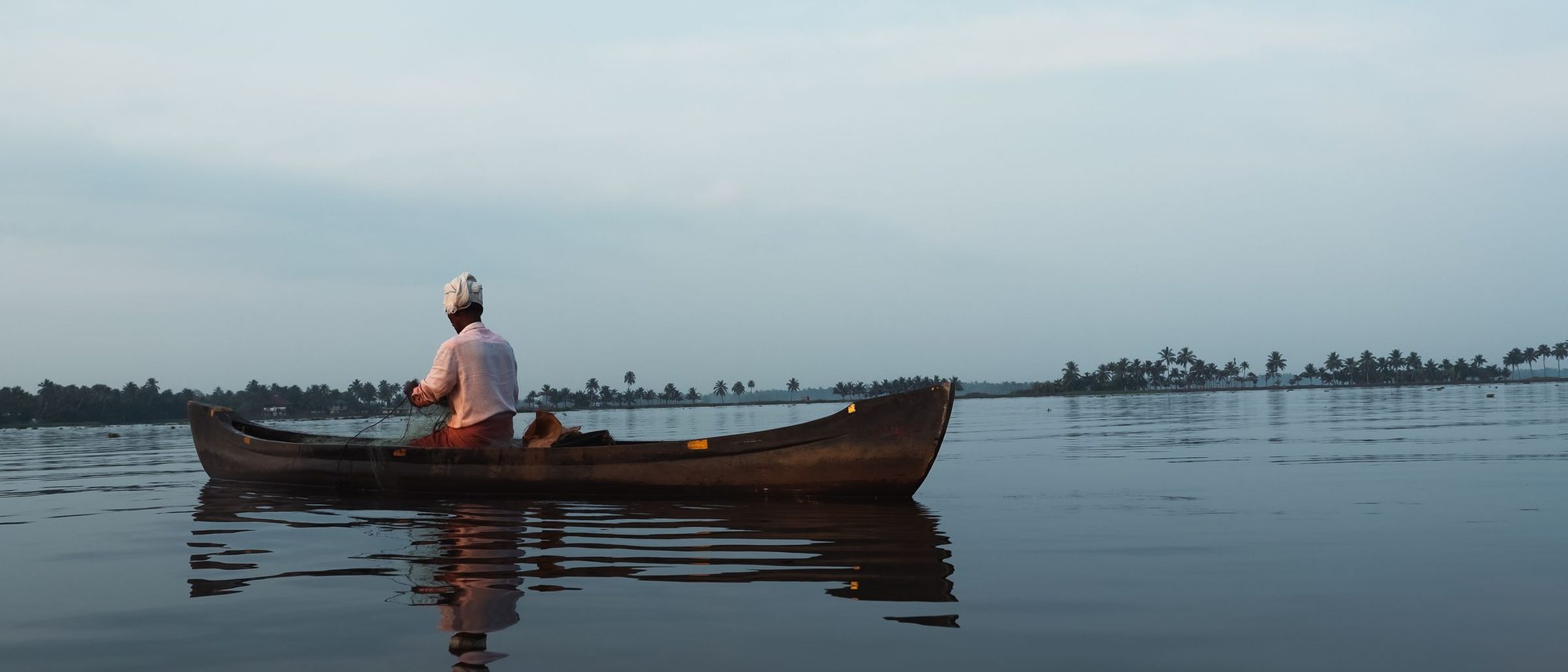 A man paddles a canoe across the still Alappuzha backwaters at dawn.