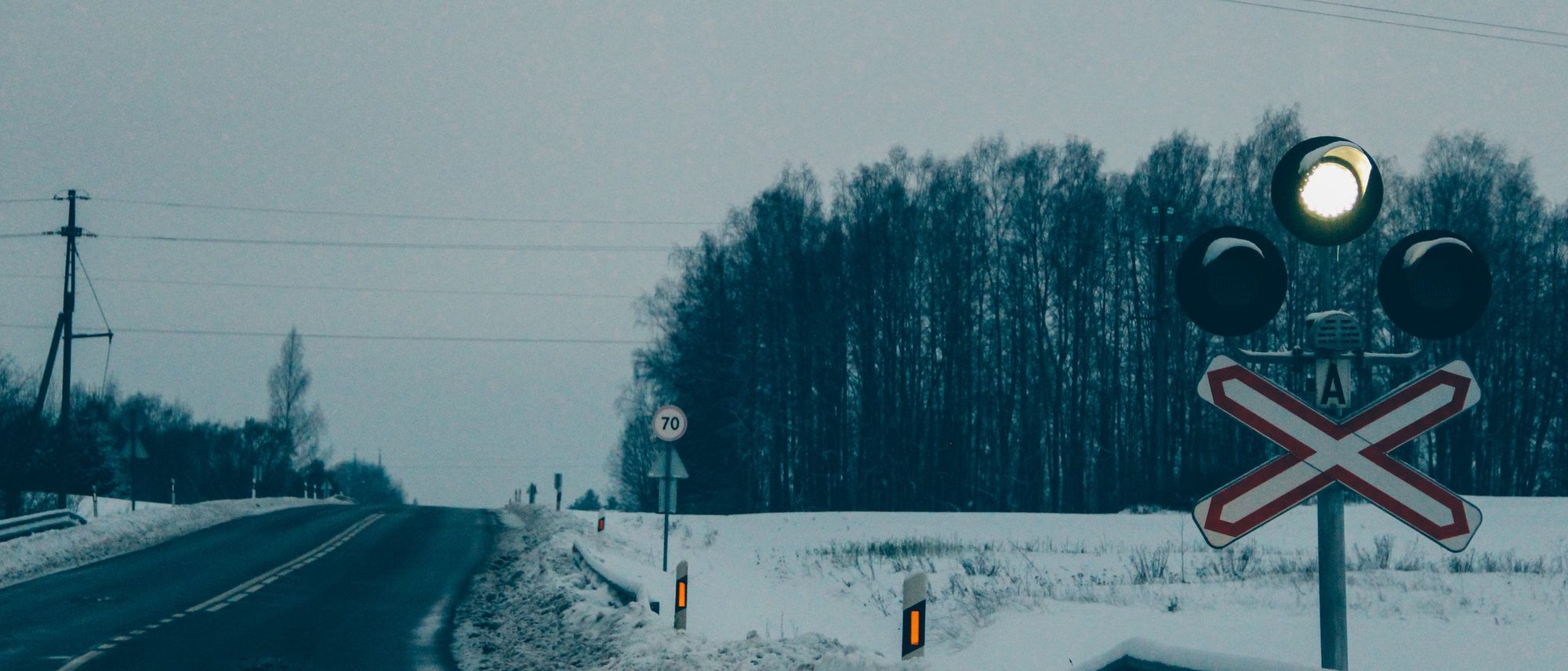A railway crossing signal beside a snowy rural road with forest behind.
