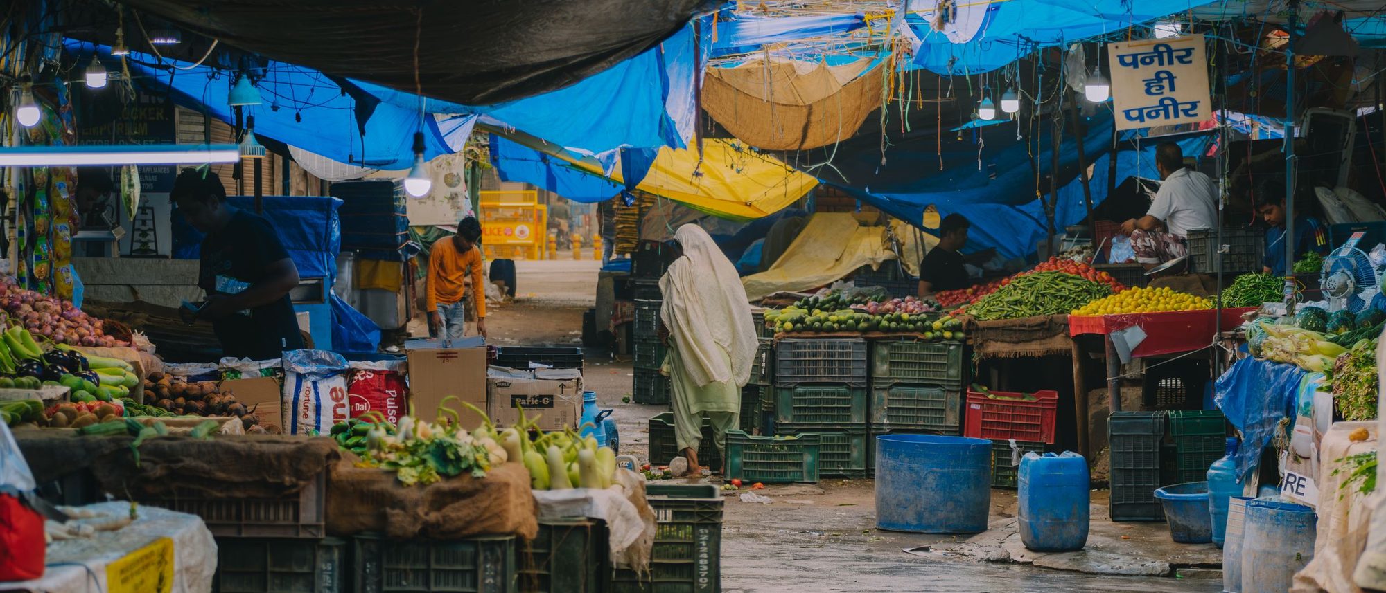 Stalls of fresh produce under coloured tarpaulins in an Indian street market on a rainy day.