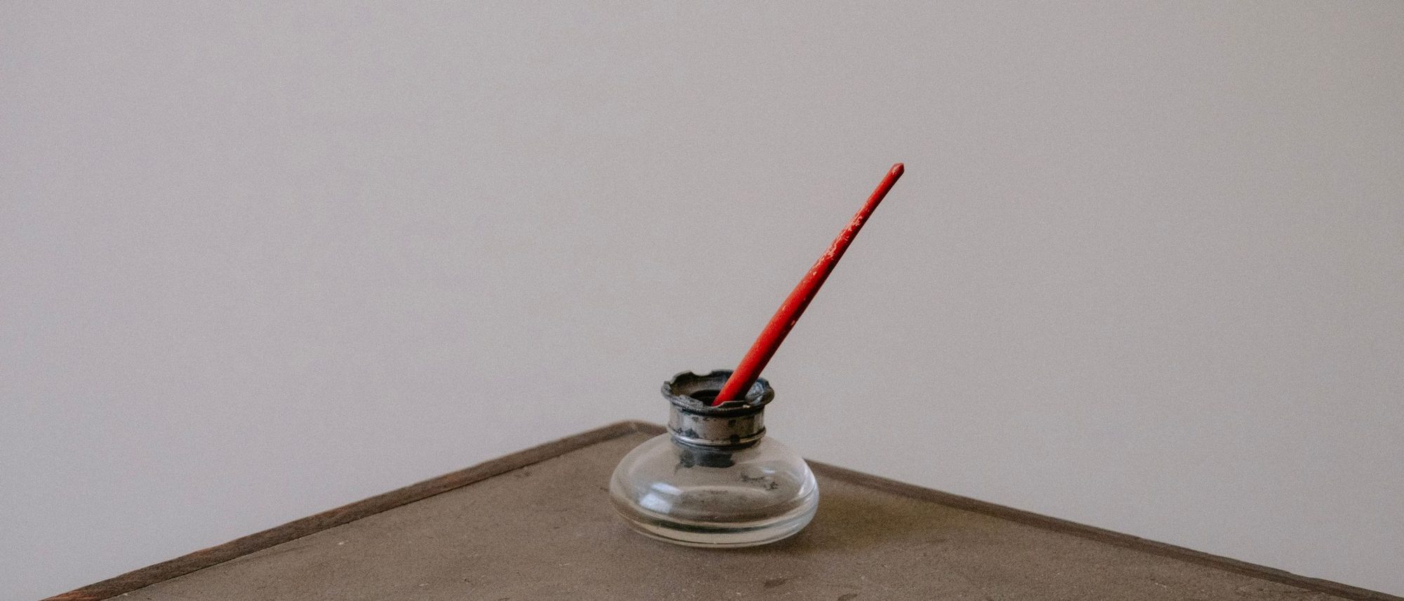 A glass inkwell with a red pen resting in it on a rustic wooden desk.